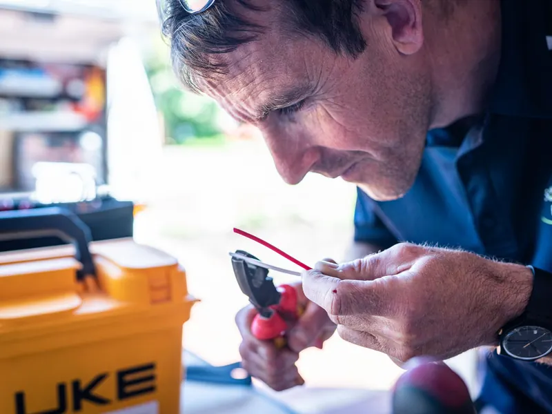 An electrician stripping a red wire beside an open tool case and a multimeter